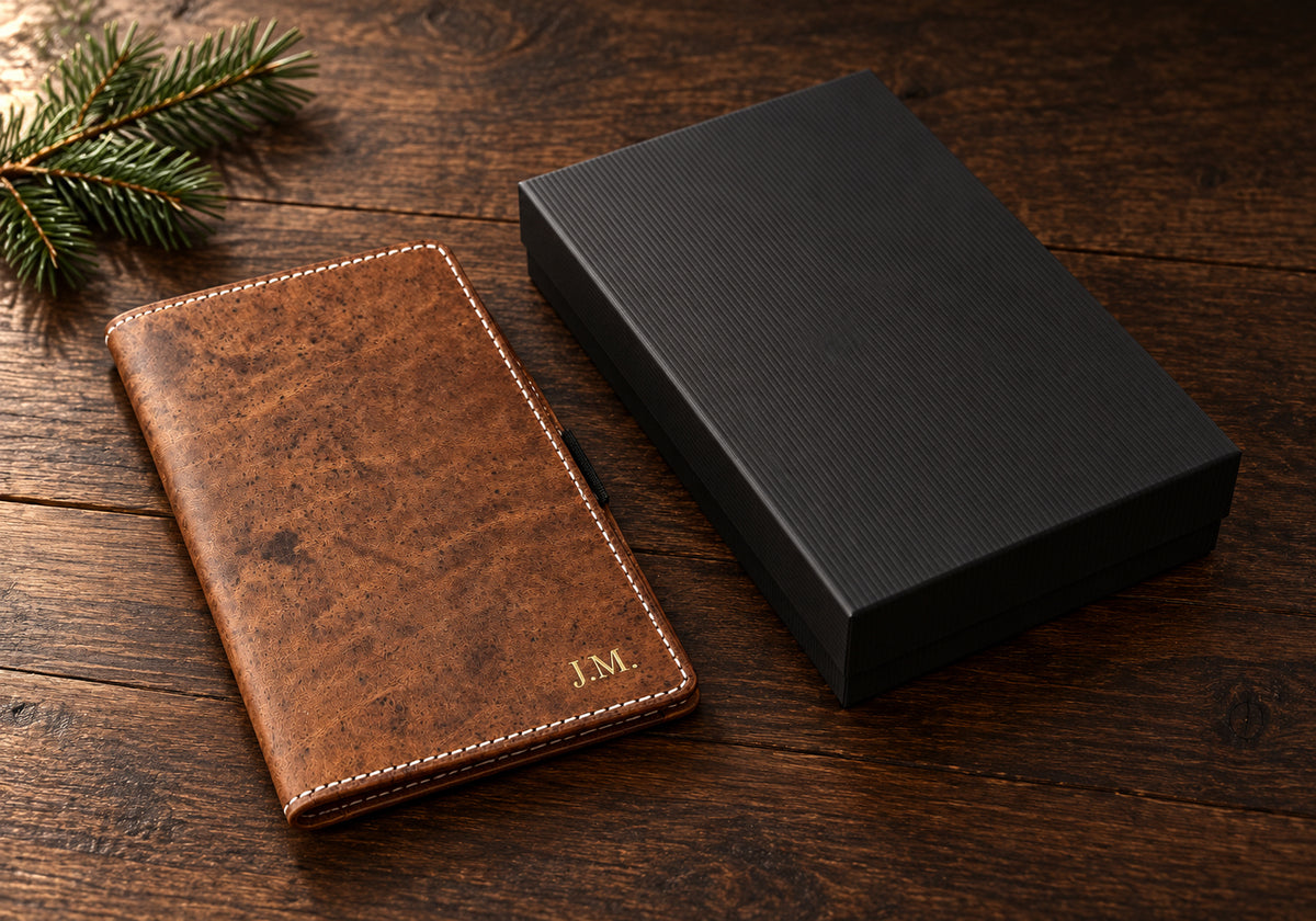 Brown leather scorecard holder with gold foil initials beside a black gift box and a sprig of pine on a walnut table