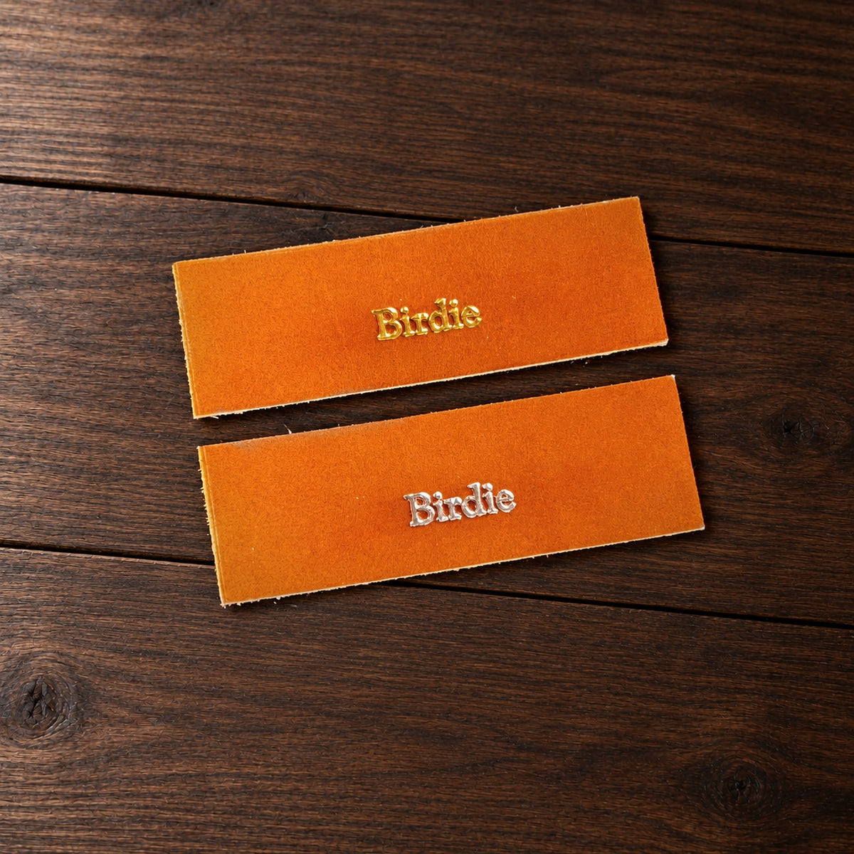 Two leather samples stamped Birdie, one in gold foil and one in silver foil, on a walnut table