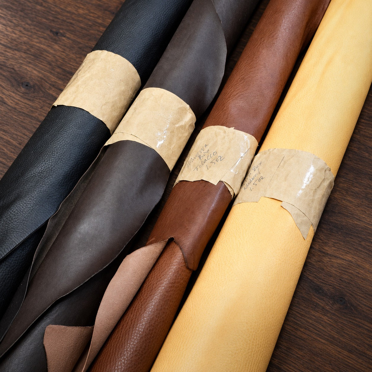 Rolls of pebbled Minerva Box leather in black, dark brown, brown and natural with kraft paper labels, on a walnut bench