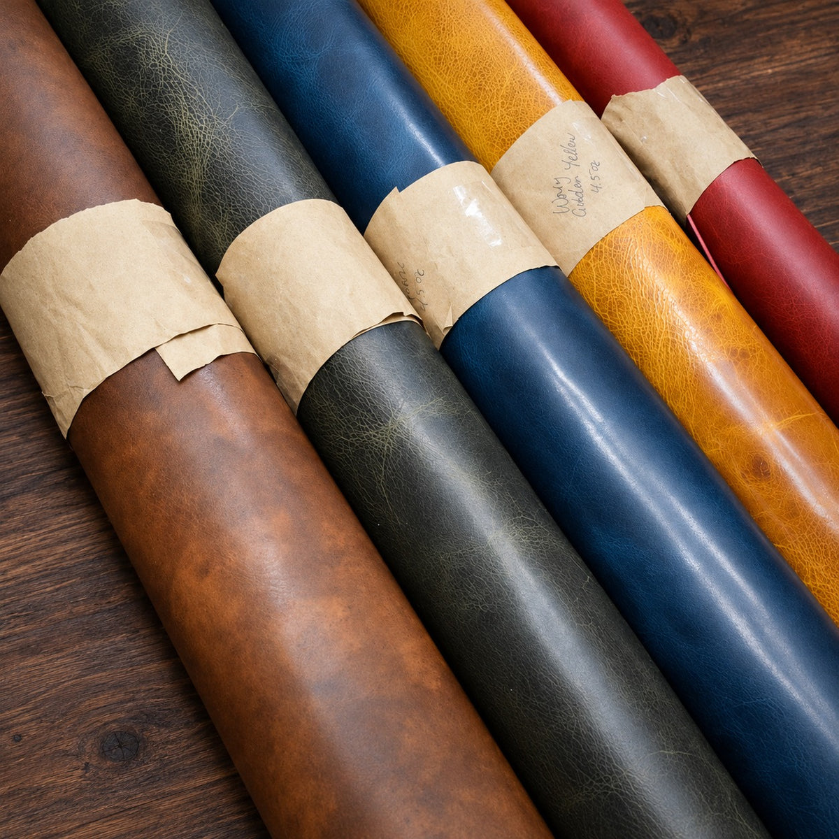 Rolls of Waxy leather in brown Nemesis, grey, blue, gold and red with kraft paper labels, on a walnut bench