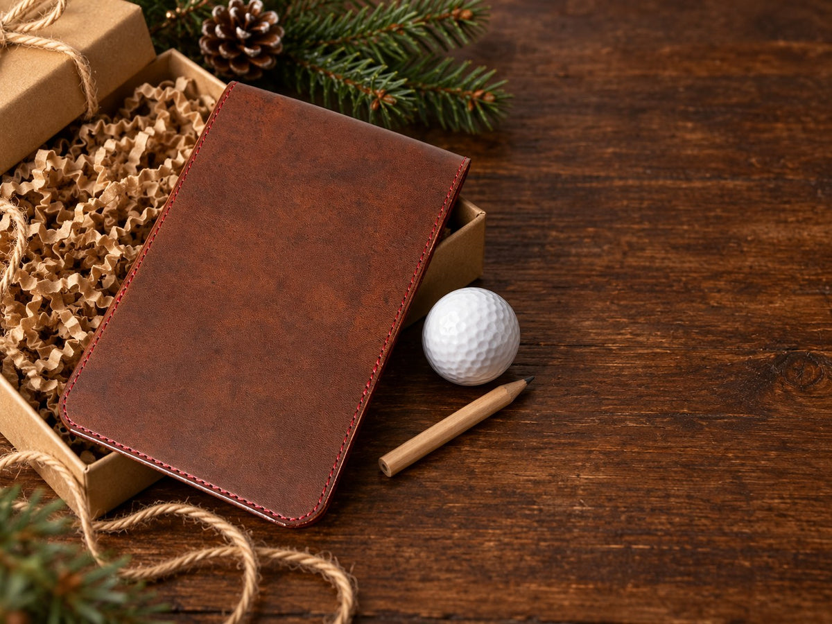 Brown leather yardage book cover with red stitching in a kraft gift box with a golf ball and pencil