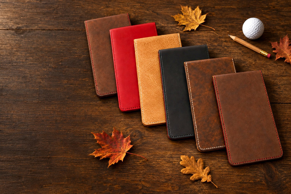 Six leather yardage book covers fanned across a walnut bench with fallen autumn leaves, a pencil and a golf ball